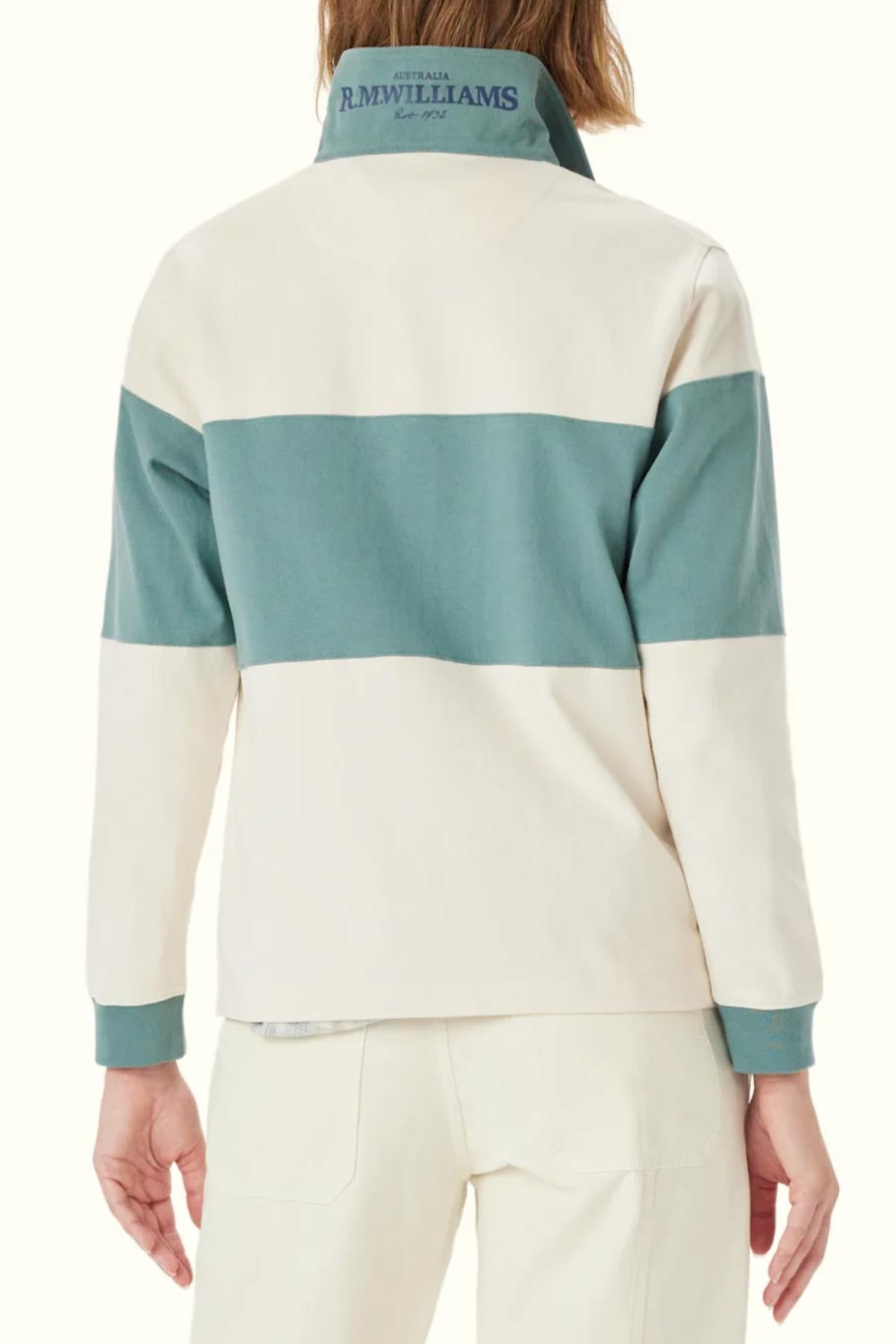 Someone wearing the RM Williams Narran Panel Stripe Rugby, a heavyweight long-sleeve shirt with wide cream and teal stripes and "R.M. Williams Australia" embroidered in blue on the collar, paired with cream pants.