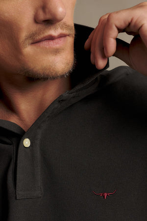 A close-up of a man wearing the RM Williams Rod Polo, featuring a black collared design with a small red bull logo embroidered on the chest. He is adjusting the collar with his hand and has light facial hair.