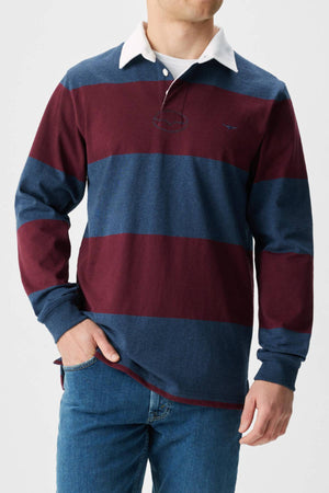 A man wearing the RM Williams Tweedale Rugby long-sleeve polo, featuring organic cotton with wide maroon and navy stripes and a white collar, pairs it with blue jeans as he stands with one hand in his pocket.