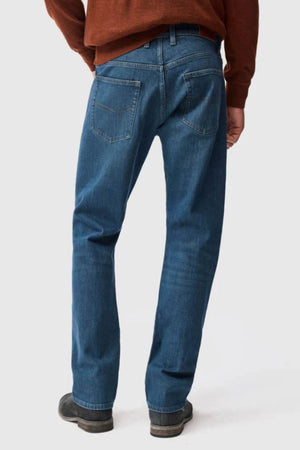 A person stands facing away, showing the back view of the Rodd & Gunn Kurow Relaxed Fit Italian Denim Jean in blue, paired with black shoes and a rust-colored long-sleeve shirt against a plain light background.