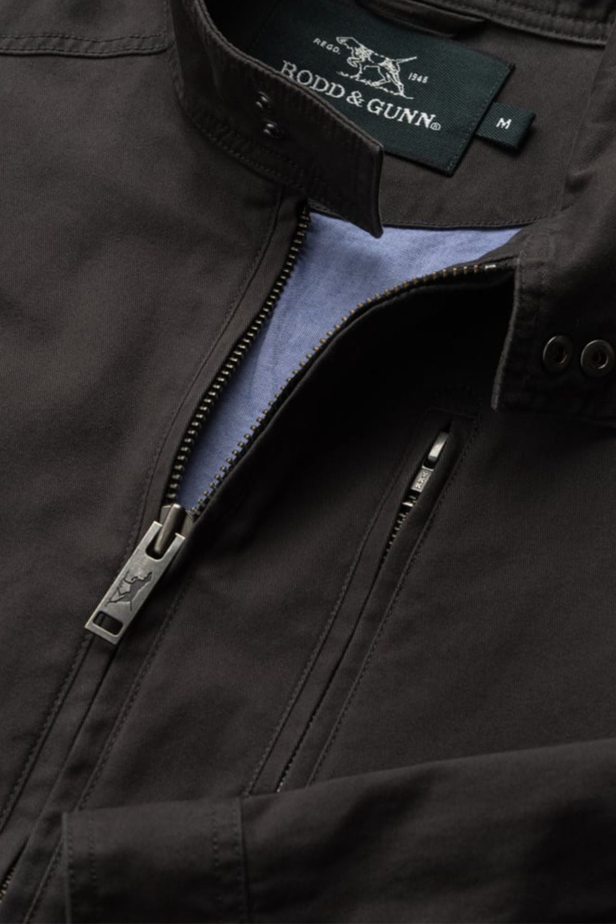 Close-up of the Rodd & Gunn Armitage Jacket in black, featuring its collar, YKK zipper with branded pull, and a partly unzipped pocket showing the light blue lining inside.