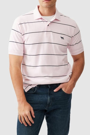 A man wears the Rodd & Gunn Glen Eden Polo, a 100% cotton light pink shirt with thin black stripes and an embroidered chest logo, paired with blue jeans. His left hand is in his pocket.