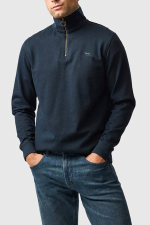 A man wears the dark navy Rodd & Gunn Gunn 14oz 1/4 Zip Sweat made with FibreTrace® Technology, standing with his hands in his pockets in blue jeans against a light gray background.