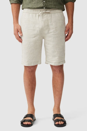 A person wearing Rodd & Gunn Linen Resort Shorts in light beige, a green rolled-sleeve long-sleeve shirt, and black sandals stands against a plain light background.