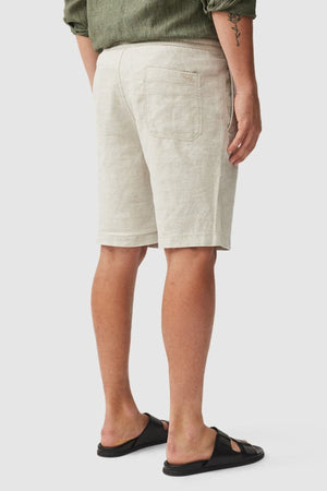 A person faces away, wearing beige Rodd & Gunn Linen Resort Shorts, a green rolled-sleeve shirt, and black slide sandals. A tattoo is visible on their left forearm. The background is plain and light-colored.