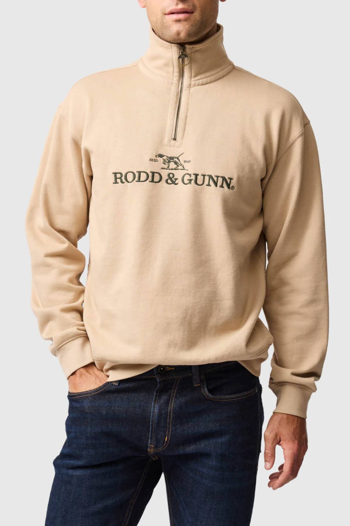 A man wears the Rodd & Gunn Milford Sound Sweat in beige, featuring "RODD & GUNN" and a bird logo on the front with a premium YKK metal zip. He pairs it with dark blue jeans and stands against a plain light gray background.