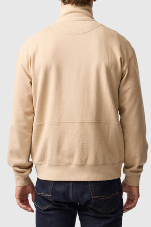 A person is seen from behind wearing a beige Rodd & Gunn Milford Sound Sweat with a premium YKK metal zip and dark blue jeans with brown stitching, set against a plain light background.