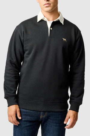 A man wearing a dark Rodd & Gunn Otago Sweat with a classic twill rugby collar and signature chest logo, paired with blue jeans, stands against a plain light gray background.