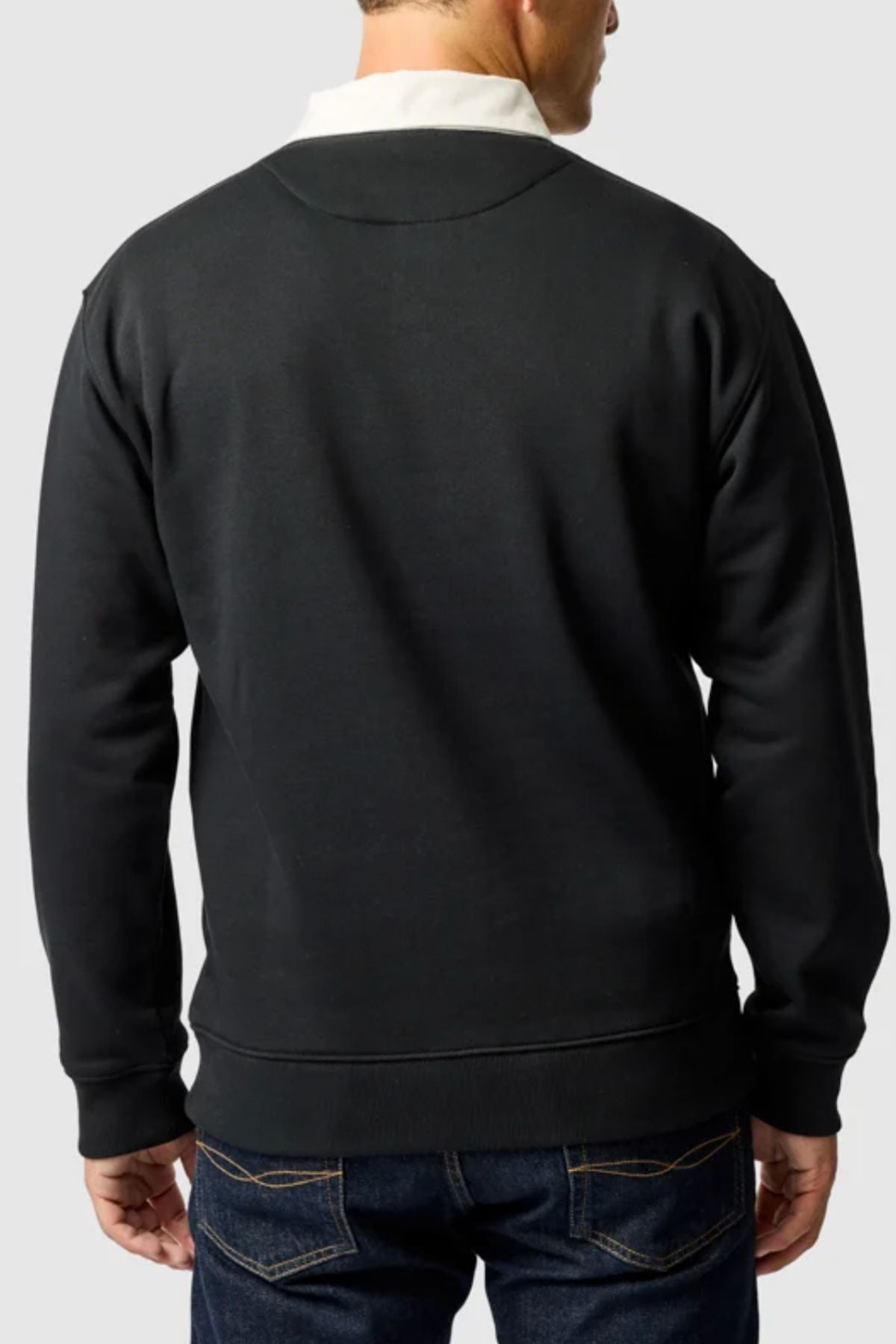 A man faces away, wearing the black Rodd & Gunn Otago Sweat featuring a traditional rugby collar over a white shirt and dark blue jeans. The subtle signature pointer logo appears on the front. The backdrop is a plain light gray.