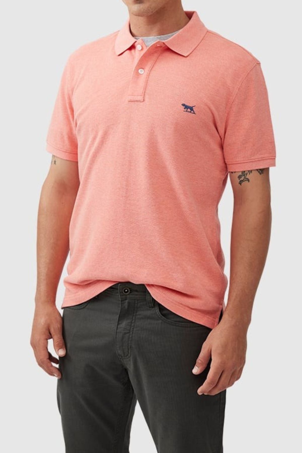 A man stands against a plain light background wearing the Rodd & Gunn The Gunn Polo—a modern fit, light pink Australian cotton shirt with a small blue embroidered chest logo—and dark gray pants, hands by his sides.