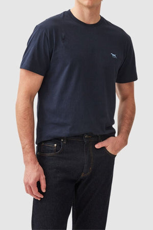 A person wearing a comfortable Rodd & Gunn The Gunn T-Shirt in navy blue, made from Australian Cotton with a small animal logo on the chest, and dark jeans. The individual is standing against a plain light gray background with their left hand in their pocket and their right arm hanging naturally by their side.