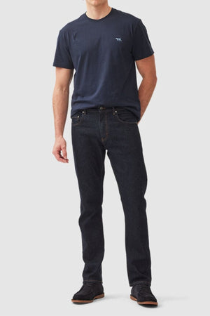 A man stands against a plain background, wearing Rodd & Gunn The Gunn T-Shirt in dark navy, made from Australian Cotton, and black straight-fit jeans. He also has on black shoes. The comfortable tee features a small white logo on the upper left side. His hands are relaxed by his sides.