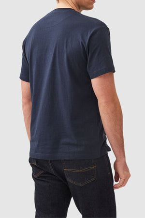 A person is standing facing away from the camera, dressed in a navy blue Rodd & Gunn The Gunn T-Shirt and dark blue jeans. The background is a plain light grey.