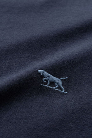 Close-up of the dark navy fabric of the Rodd & Gunn The Gunn T-Shirt, featuring an embroidered design of a small, light blue dog. The slightly textured material displays the dog in a standing or walking position, emphasizing a subtle and minimalist style, ideal for this comfortable tee made from Australian Cotton.