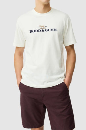 Wearing a white Rodd & Gunn The Peaks Logo Tee and dark brown shorts, a person stands with hands in pockets against a plain light grey background.