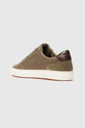 The Rodd & Gunn Transit Sneaker features an olive green upper, white sole, brown leather heel accent, suede details, and a translucent outsole. Shown from a rear-side angle on a plain light grey background.