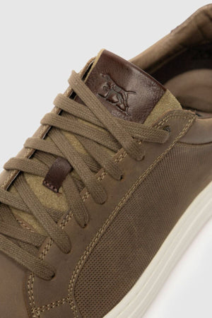 Close-up of the Rodd & Gunn Transit Sneaker featuring a textured leather upper, matching brown laces, a dark brown tongue with an embossed polo player logo, and a white translucent outsole.