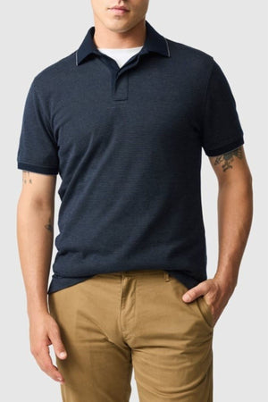 A man wearing the Rodd & Gunn Wilson Bay Sports Fit Polo in navy and tan pants stands against a plain light gray background, one hand in his pocket, with his face partially out of frame.