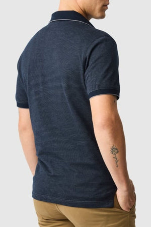A man stands with his back to the camera in a navy Rodd & Gunn Wilson Bay Sports Fit Polo made from breathable birdseye knit and khaki pants. A small plant or flower tattoo is visible on his left forearm.