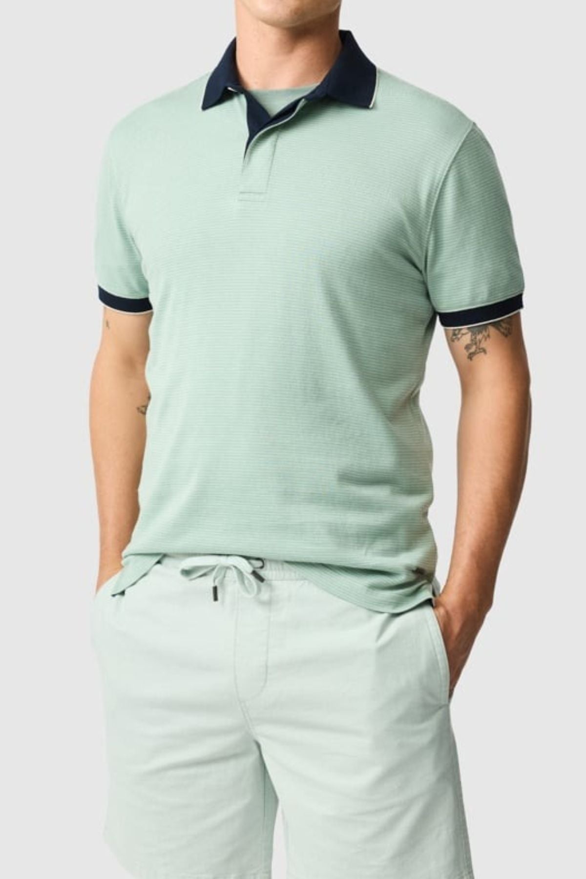 A person wearing a Rodd & Gunn Wilson Bay Sports Fit Polo in light green with dark green accents, paired with matching shorts. The breathable birdseye knit offers comfort as hands rest in pockets. Tattoos are visible on the arms against a plain gray background.