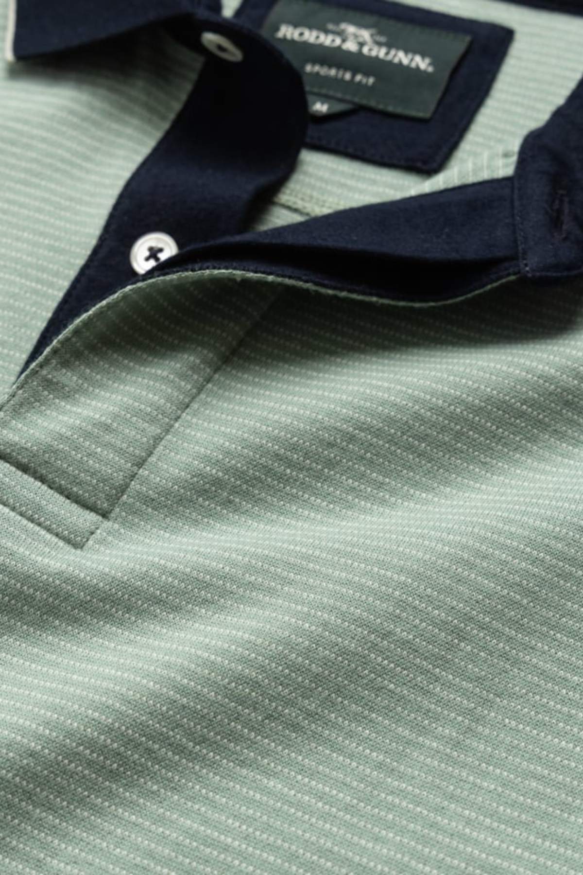 The Rodd & Gunn Wilson Bay Sports Fit Polo showcases a green, breathable birdseye knit with a subtle dotted pattern and a contrasting dark navy collar. Near the neckline are three buttons, while the back label reads "Rodd & Gunn.