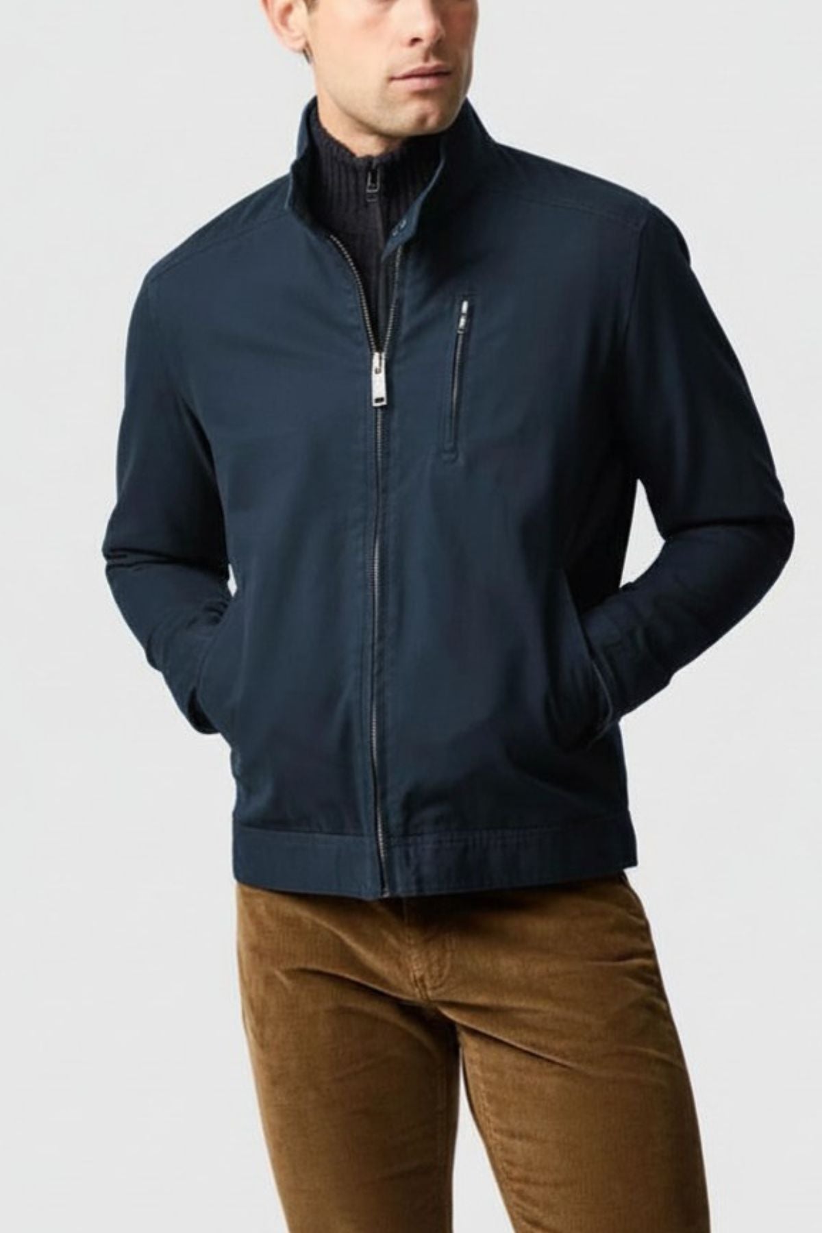 A man stands against a plain light gray background, hands in pockets, wearing a navy Rodd & Gunn Armitage Jacket over a black top and brown corduroy pants.