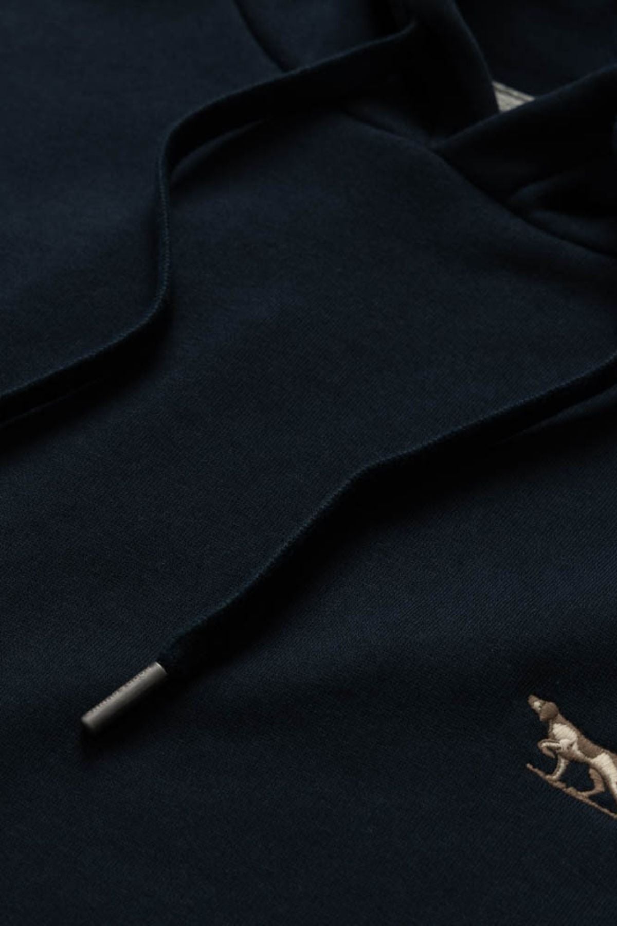 Close-up of the Rodd & Gunn Otago Hoodie, crafted from dark Australian cotton and featuring black drawstrings with a small embroidered dog logo in the lower right corner, highlighting Rodd & Gunnβs signature attention to detail.