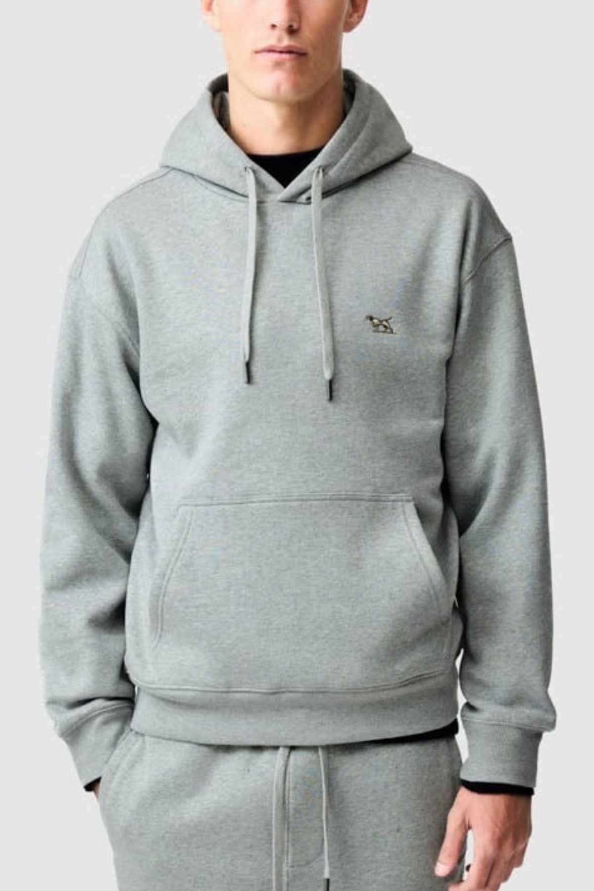 Wearing the Rodd & Gunn Otago Hoodie in light gray, a person stands with hands in pockets against a plain light background, dressed in matching Australian Cotton brushed back fleece sweatpants.
