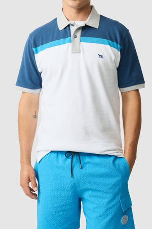 A person wears the Rodd & Gunn Whitby Polo with a color block design in white, blue, and gray, paired with bright blue drawstring shorts. They pose with one hand in their pocket against a plain light background.