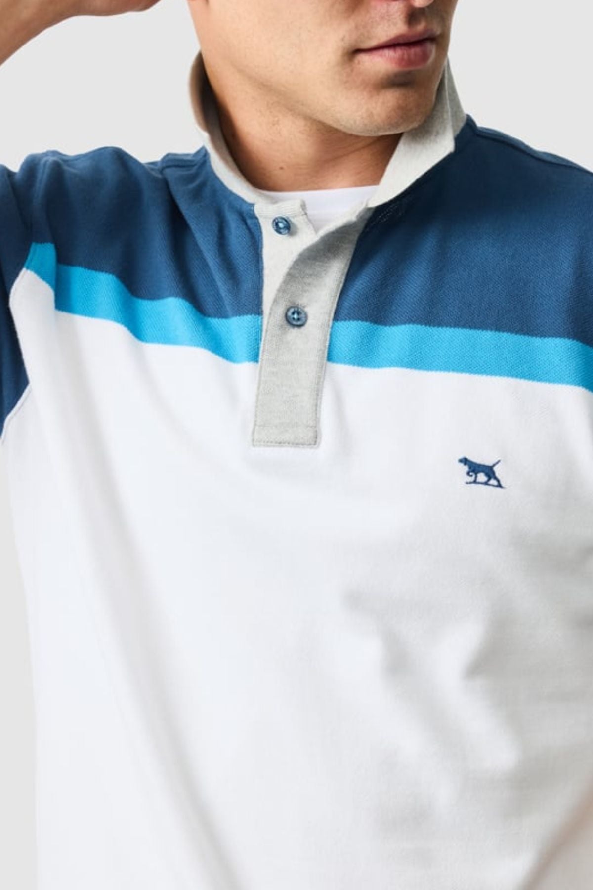 A man wears a Rodd & Gunn Whitby Polo featuring a white and blue color block design, light gray placket, blue buttons, a chest stripe, and an embroidered animal logo on the left chest. His face is partially visible.