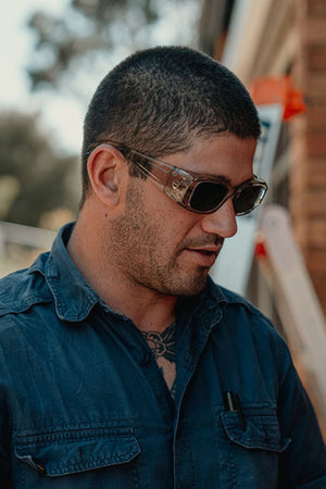 A man with short dark hair wears SafeStyle Auras Graphite Frame Grey Polarised Lens sunglasses and a blue button-up shirt, standing outdoors by a building and looking down with a neutral expression.
