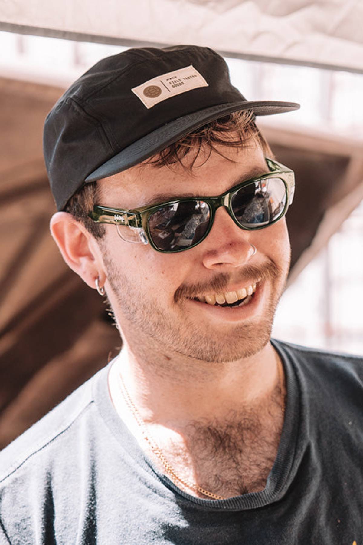 A man wearing SafeStyle Classics Green Frame Polarised Lens sunglasses, a black cap, navy t-shirt, and chest chain smiles outdoors. He has a mustache, light facial hair, and sunlight highlights his face.