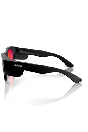 SafeStyle Cruisers Sunglasses feature matte black frames and mirror red polarized lenses in a side view. The temple arm displays a small white logo, providing UV protection, with their stylish design casting a reflection on the white surface below.