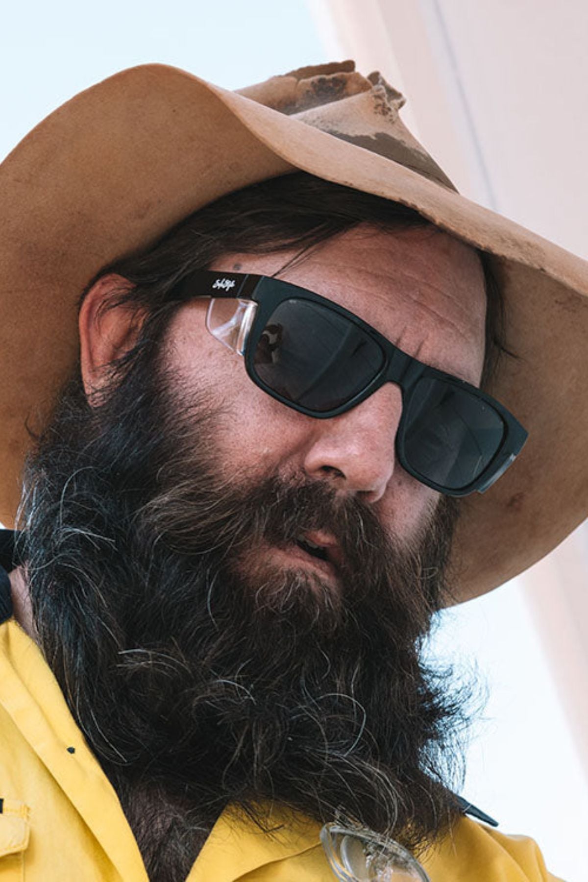 A bearded man in a wide-brimmed hat and a yellow shirt with a brown collar gazes downward. He sports SafeStyle Fusions XL Black Frame Polarised Lens sunglasses, ideal for sunny outdoor settings, subtly highlighting his stylish eyewear choice against the blurred background.