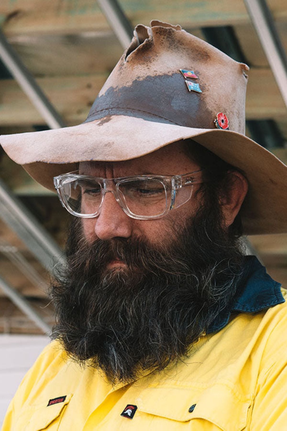 A man with a large beard wears SafeStyle Fusions XL Clear Frame glasses and a worn, pin-adorned wide-brimmed hat. Dressed in a yellow and blue shirt, he stands thoughtfully in front of a structure of metal beams and wood.