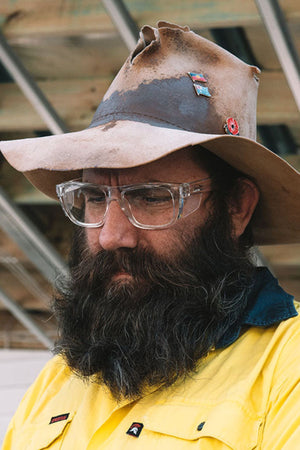 A man with a large beard wears SafeStyle Fusions XL Clear Frame glasses and a worn, pin-adorned wide-brimmed hat. Dressed in a yellow and blue shirt, he stands thoughtfully in front of a structure of metal beams and wood.