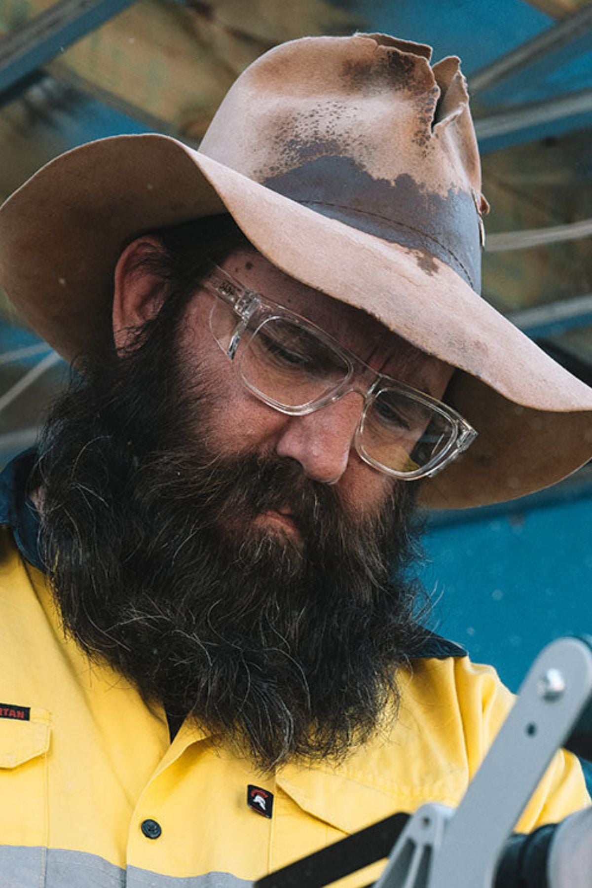 A person with a long beard, wearing a wide-brimmed hat and SafeStyle Fusions XL clear frame safety glasses, is focused on something off-camera. They are dressed in a yellow and dark blue work shirt against an industrial background.