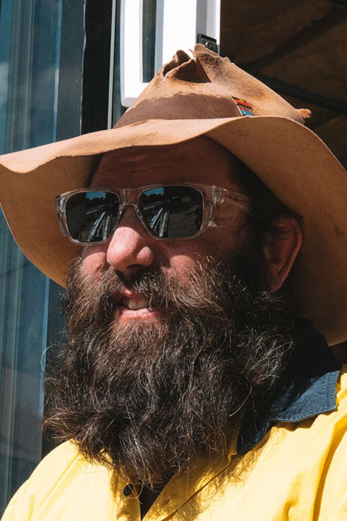 A bearded person in a yellow shirt and old brown hat stands outside in bright sunlight, wearing SafeStyle Fusions XL Clear Frame Polarised Lens sunglasses; light reflects off a glass window behind them.