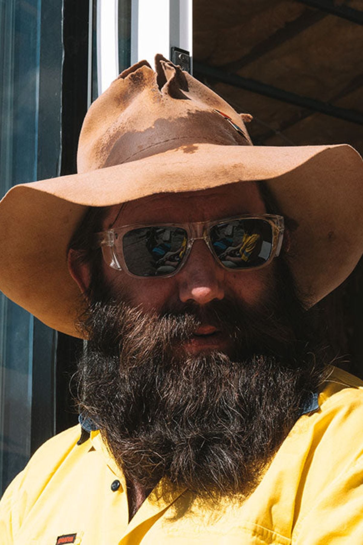A man with a thick dark beard wears a wide-brimmed brown hat, a bright yellow shirt, and SafeStyle Fusions XL Clear Frame Polarised Lens sunglasses while standing outdoors in bright sunlight.