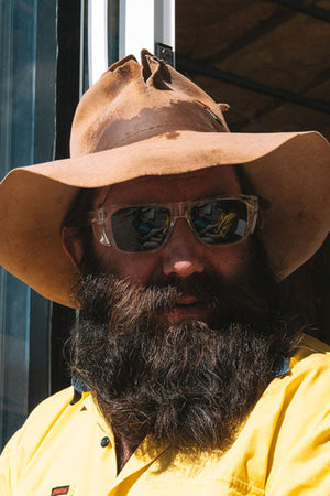 A man with a thick dark beard wears a wide-brimmed brown hat, a bright yellow shirt, and SafeStyle Fusions XL Clear Frame Polarised Lens sunglasses while standing outdoors in bright sunlight.