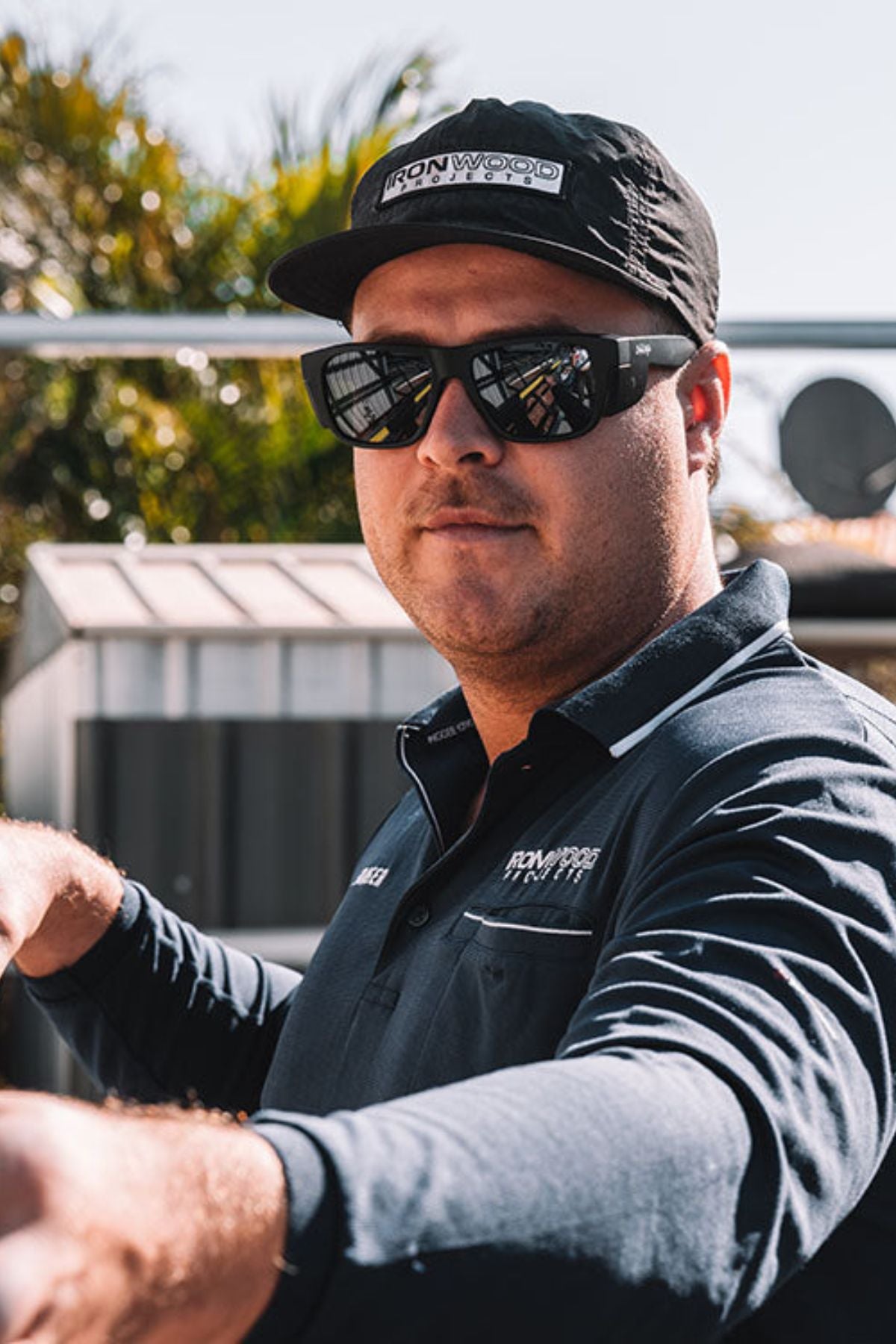 A man sporting the SafeStyle Fusions XL Matte Black Frame Polarised Lens sunglasses, along with a navy blue polo shirt and a black cap, stands outdoors on a sunny day. He gazes at the camera with a neutral expression, set against a backdrop of greenery and part of a building.