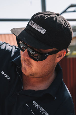 A man, wearing a black SafeStyle Fusions XL Matte Black Frame with polarised lenses for UV protection, is clad in a navy-blue shirt emblazoned with "Ironwood Projects" and "Fraser." He seems focused, looking down and possibly working on something outdoors, suggesting his involvement in construction or similar tasks.