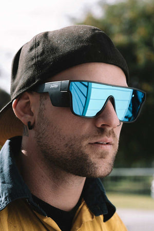 A person wearing a black cap backwards, large SafeStyle Primes Matte Black Frame Reflectors Blue Lens sunglasses, and a yellow and black jacket stands outdoors. The background is blurred, with hints of greenery.