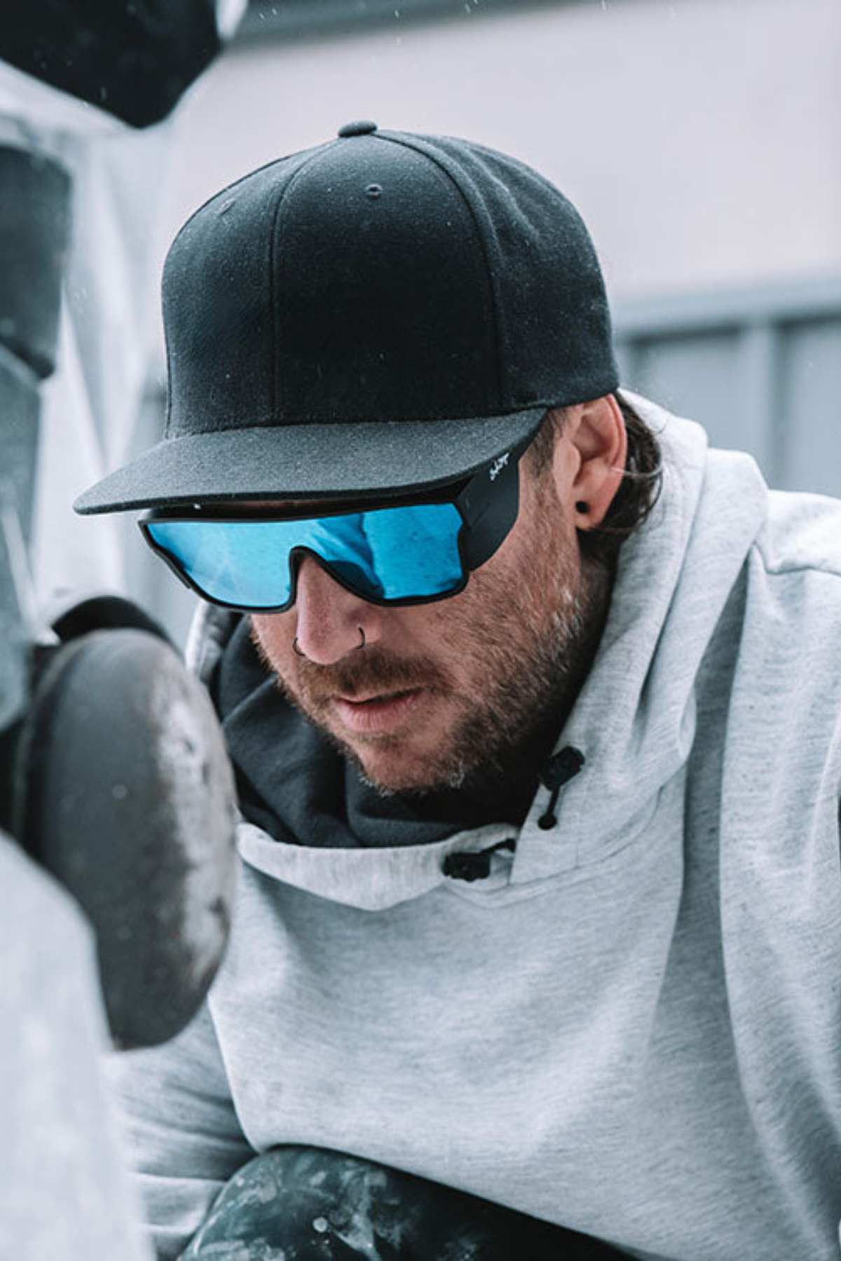 A person wearing SafeStyle Primes Matte Black Frame Reflectors with Blue Lens and UV protection, along with a black cap, mirrored sunglasses, and a gray hoodie, is closely observing something out of view. The background appears to be an outdoor setting with industrial elements.