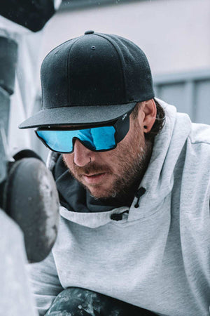 A person wearing SafeStyle Primes Matte Black Frame Reflectors with Blue Lens and UV protection, along with a black cap, mirrored sunglasses, and a gray hoodie, is closely observing something out of view. The background appears to be an outdoor setting with industrial elements.