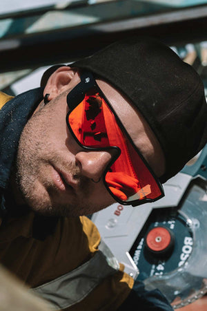 A man wearing SafeStyle Primes Matte Black Frame Reflectors Red Lens sunglasses and a black cap backwards looks at the camera. He has a light beard, wears a yellow and dark jacket, with machinery featuring buttons and dials in the background.