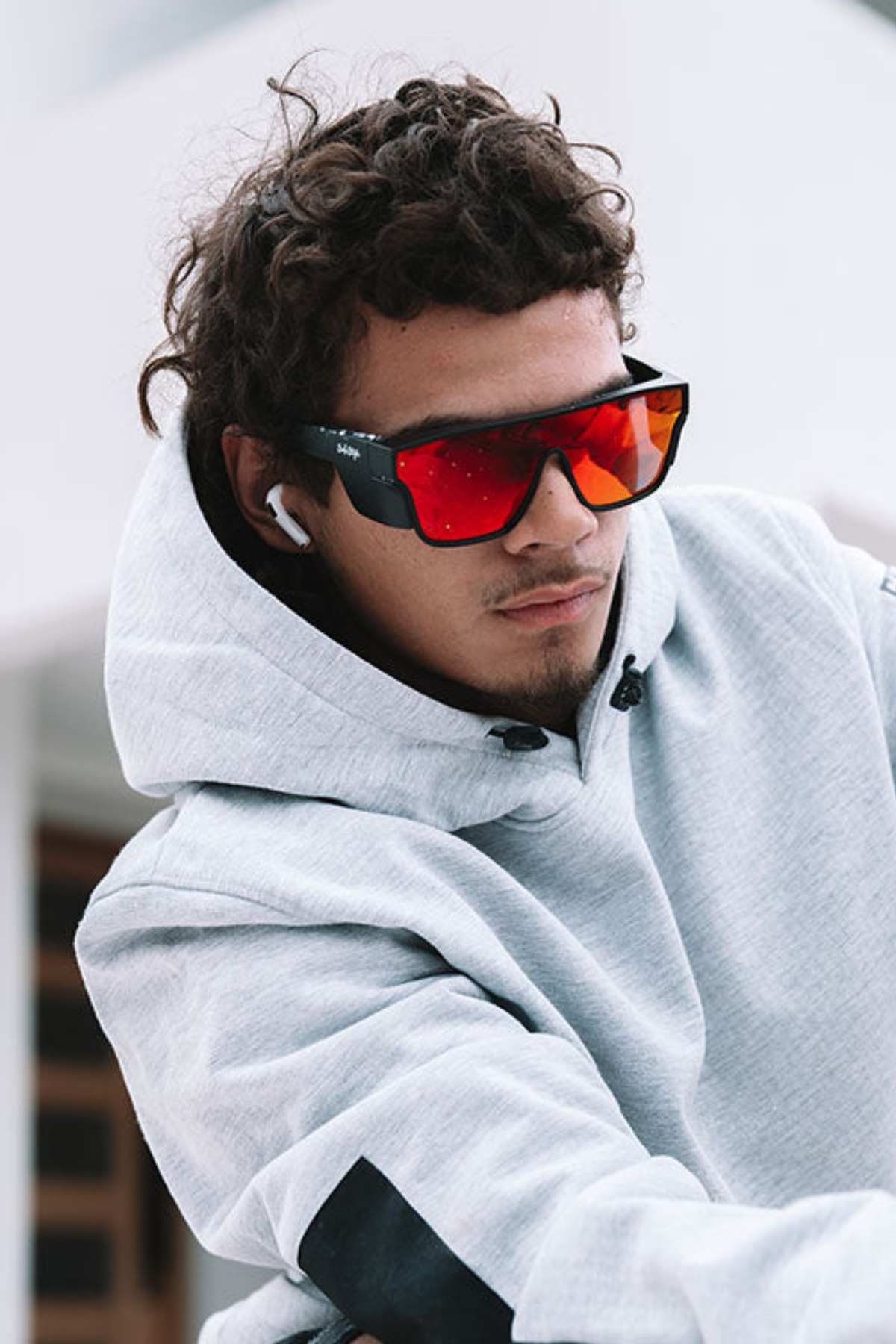 A young man with curly hair wears a light gray hoodie and SafeStyle Primes Matte Black Frame Reflectors Red Lens sunglasses with UV protection, plus wireless earbuds, looking down seriously against a blurred outdoor background.
