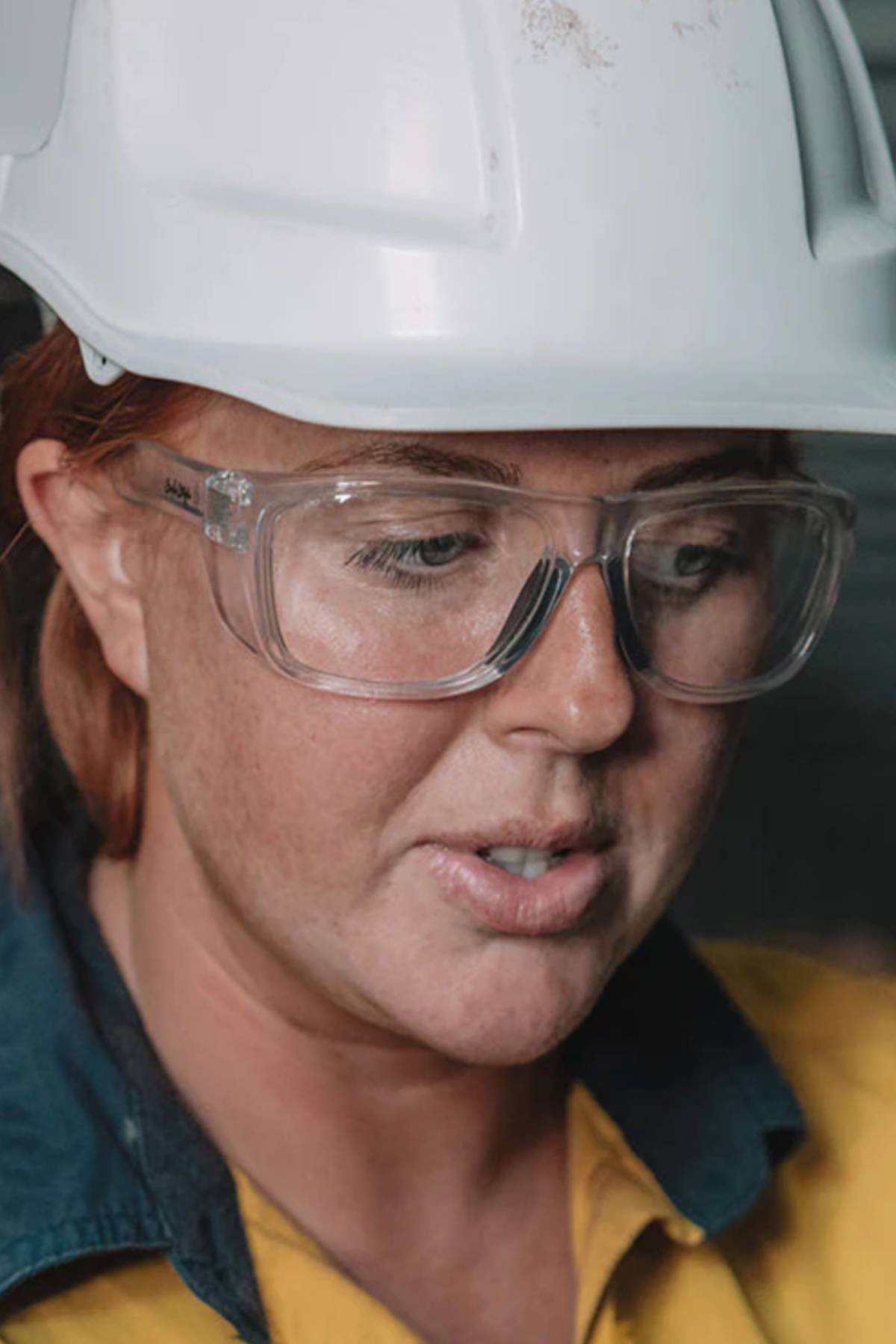 A close-up of a person wearing a white hard hat, yellow shirt, and SafeStyle Rapids Clear Frame Clear Lens safety glasses, featuring shatterproof frame and lenses, looking downward with a focused expression.