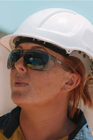 A person with light skin and reddish hair in a collared work shirt wears a white helmet and SafeStyle Rapids Graphite Frame Polarised Lens sunglasses, looking to the side, possibly at a construction site.