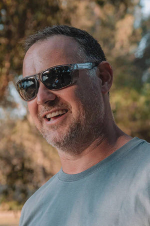 A man smiles outdoors wearing SafeStyle Rapids Graphite Frame Polarised Lens sunglasses and a grey t-shirt, with blurred trees and sunlight in the background.